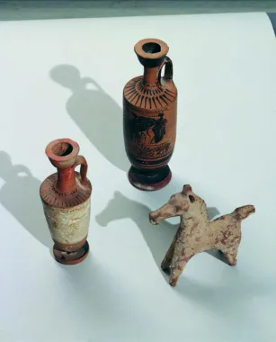 Tre græske genstande, tv. en hvid lekythos, en salveflaske, højde 16,7 cm. Bagerst en sort lekythos, højde 20,8 cm, begge lekyther er attiske, ca. 500-400 f.Kr. Til højre en lille hest i arkaisk stil, ca. 600-480 f.Kr., længde 13 cm.